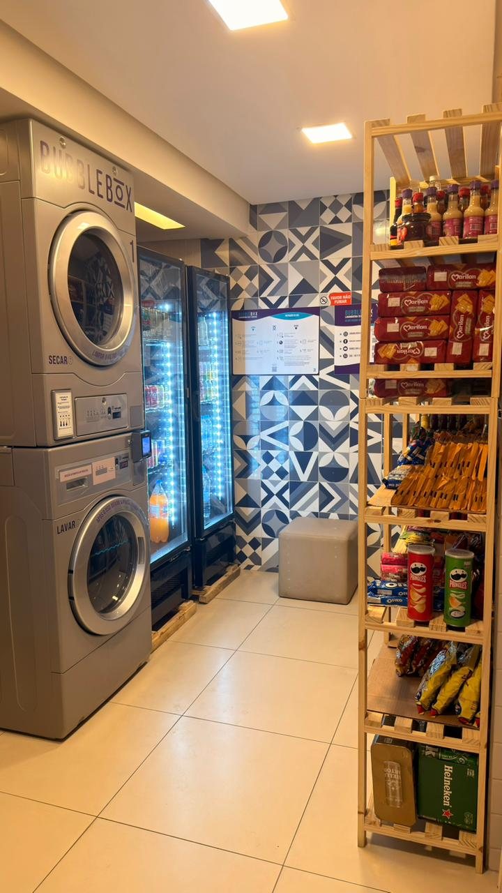 Vista de quem chega: lavadoras à esquerda, geladeiras de bebidas ao fundo diante da parede de azulejos e prateleiras de madeira cheias de produtos à direita.