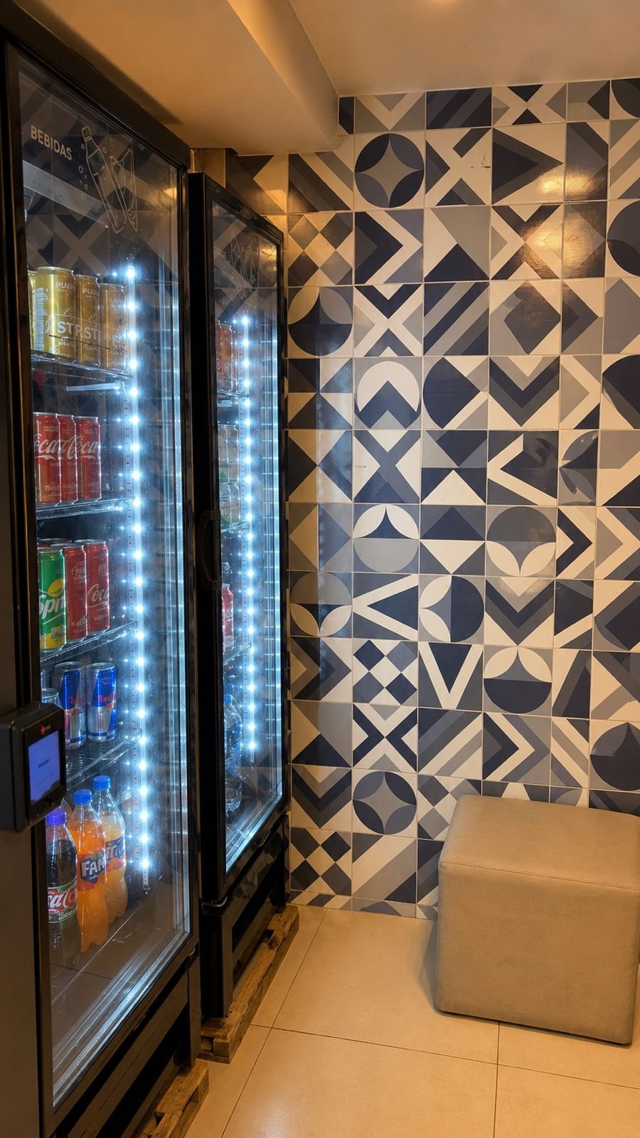 Duas geladeiras de porta de vidro iluminadas com latas de refrigerante, energéticos, sucos e garrafas, ao lado da parede de azulejos geométricos azuis.