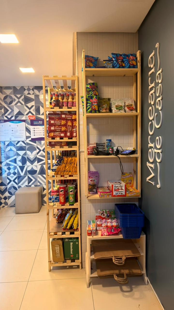 Prateleiras de pinus com salgadinhos, batatas, biscoitos, doces e chocolates, ao lado da parede azul com o letreiro Mercearia de Casa.
