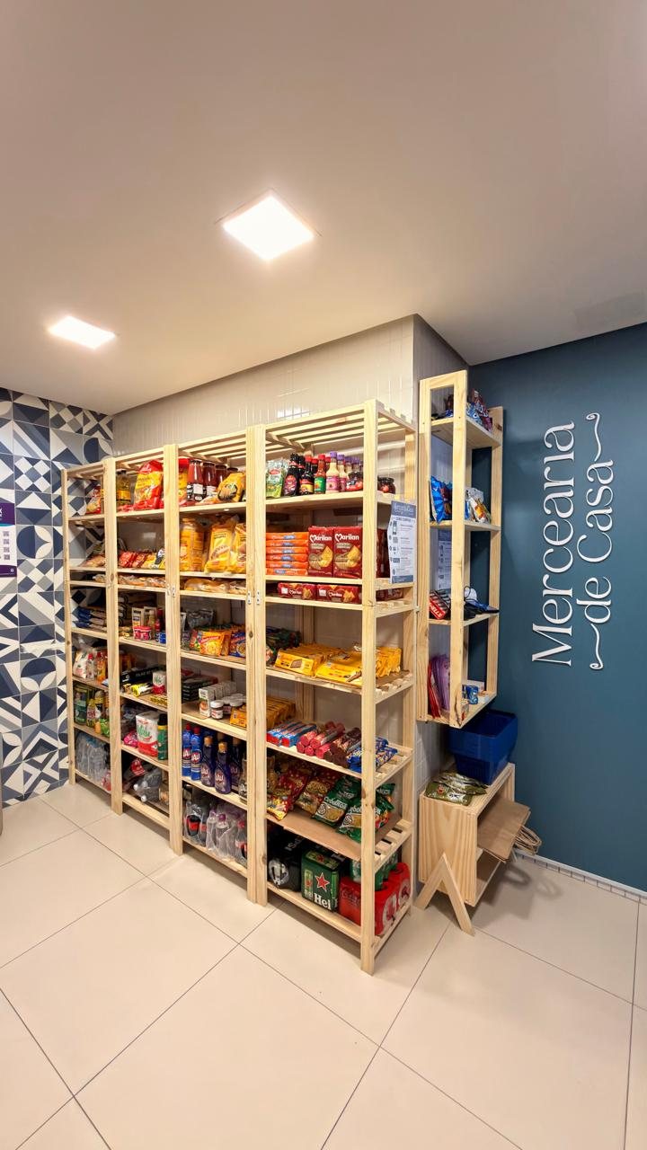 Estantes de pinus do chão ao teto abastecidas com molhos, biscoitos, massas, produtos de limpeza e bebidas, com a logo Mercearia de Casa na parede azul.