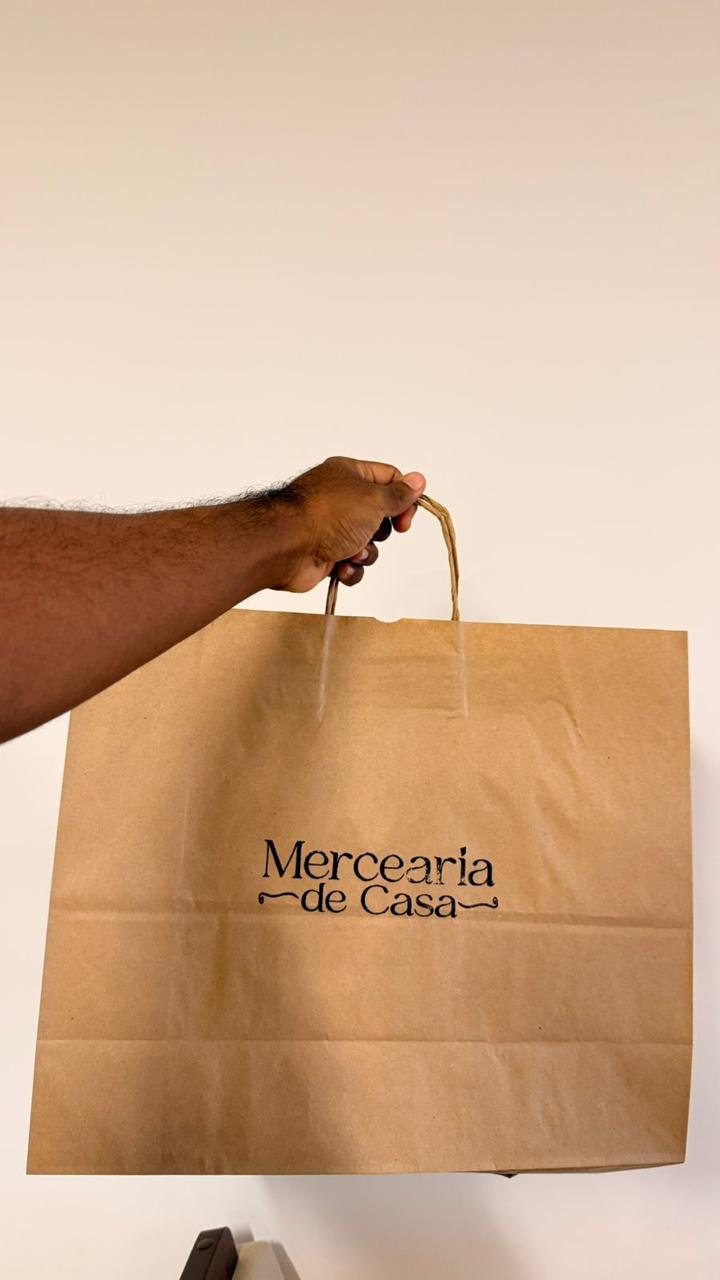 Mão segurando uma sacola de papel kraft com a logo Mercearia de Casa.
